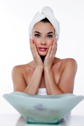 Woman wearing towel and hair in towel touching sides of face in front of spa water bowl